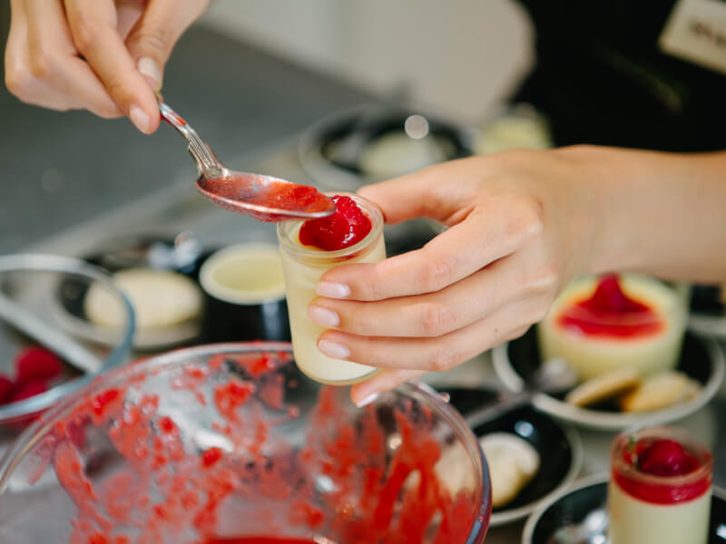 A close up of a hand spreading red puree on to the top of a cream dessert